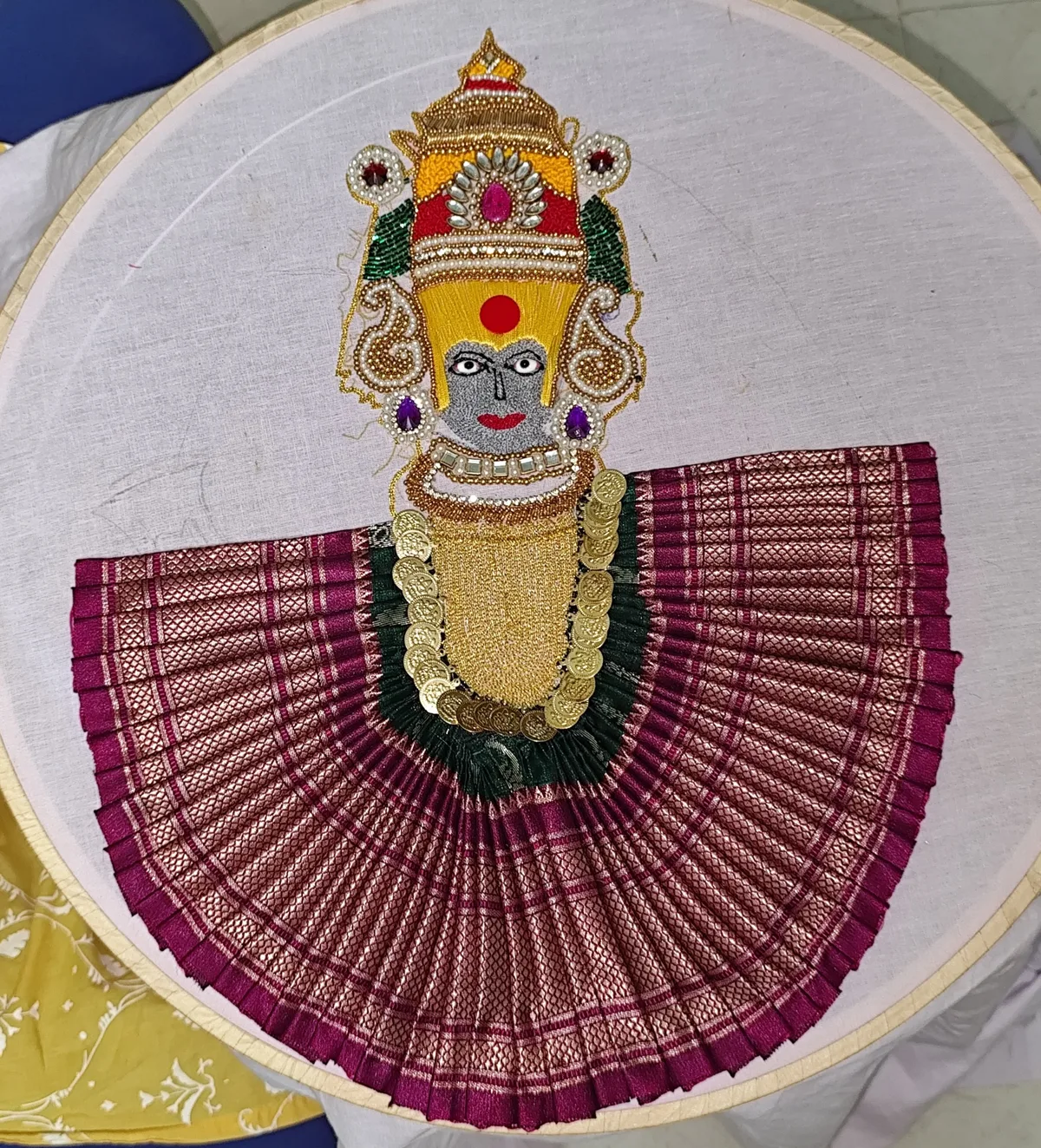 Aari embroidery of a traditional deity with gold beadwork, pearls and a pleated magenta fabric skirt, on an embroidery hoop